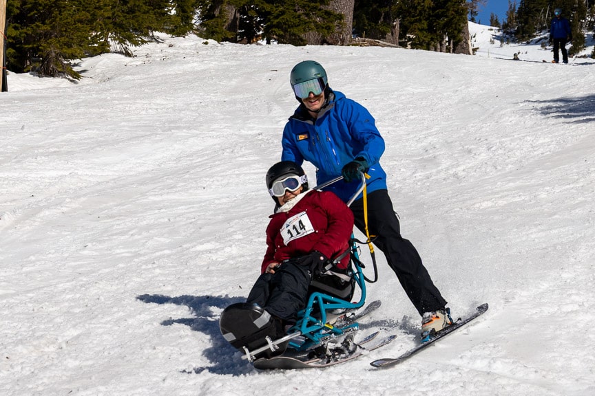 A person on a sit-ski with a person on skis go down the mountain